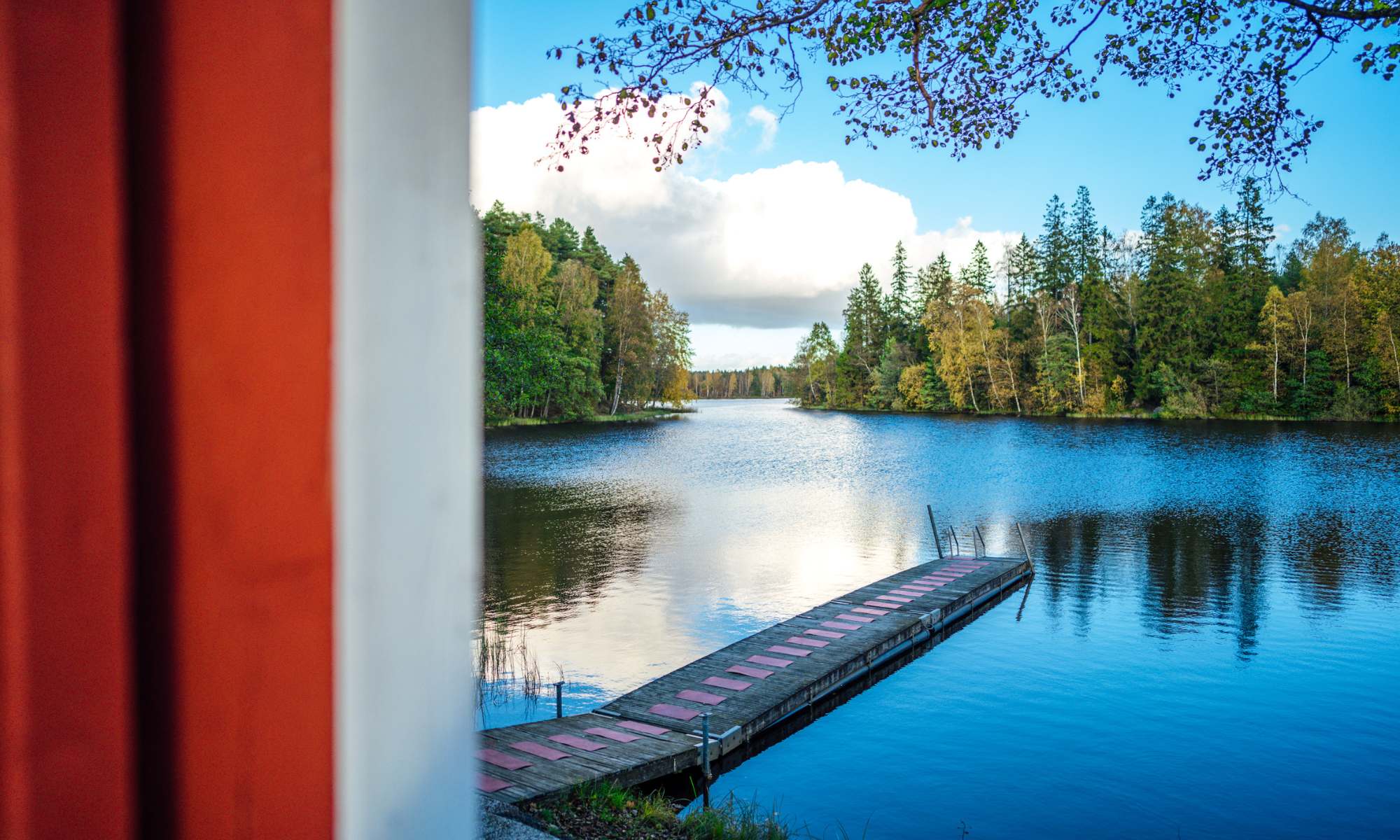 Utsikt över en sjö med en brygga i och man ser lite av en röd husknut på bastun