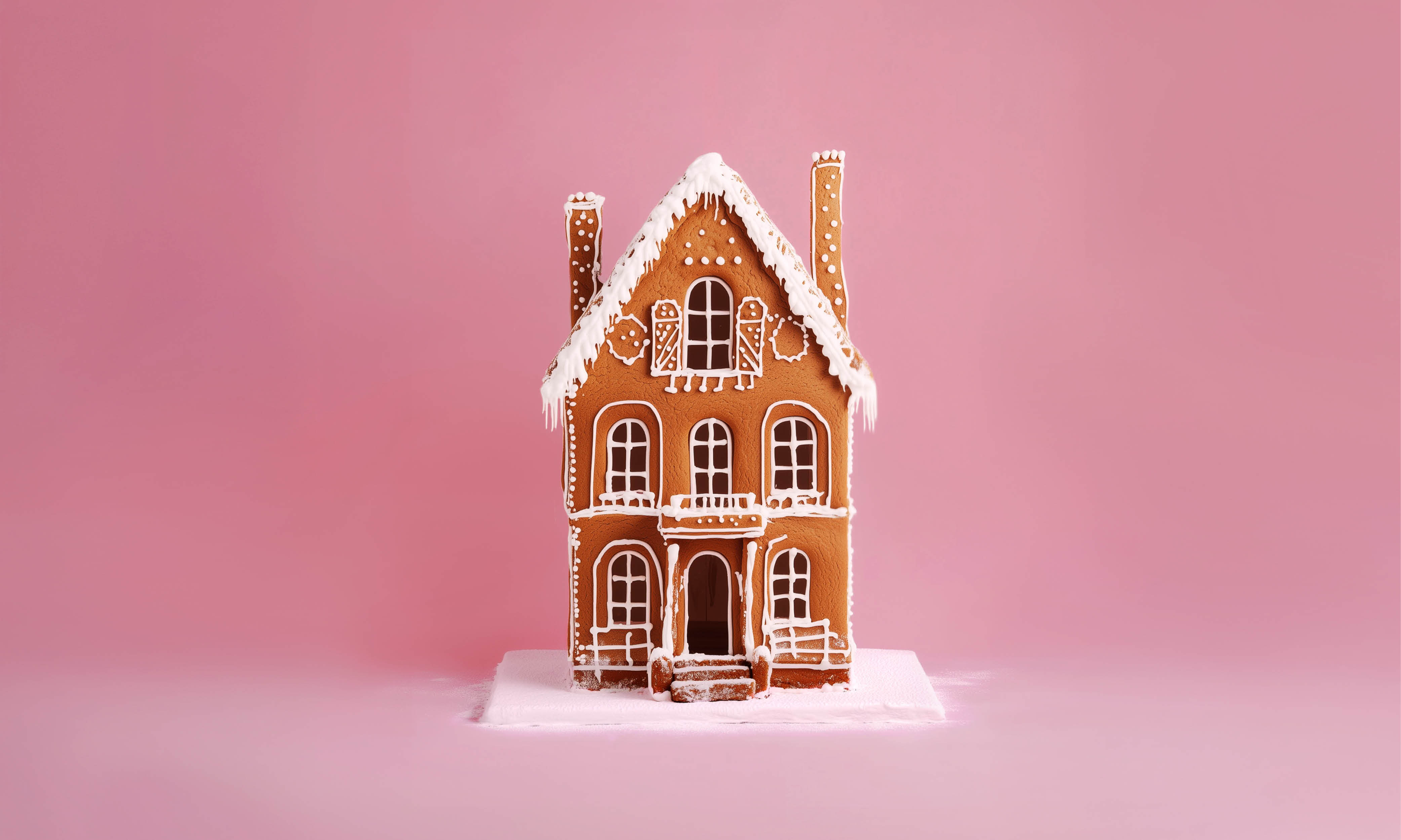 Ett detaljerat pepparkakshus dekorerat med vit glasyr står mot en rosa bakgrund, med intrikata mönster och ett snötäckt tak.