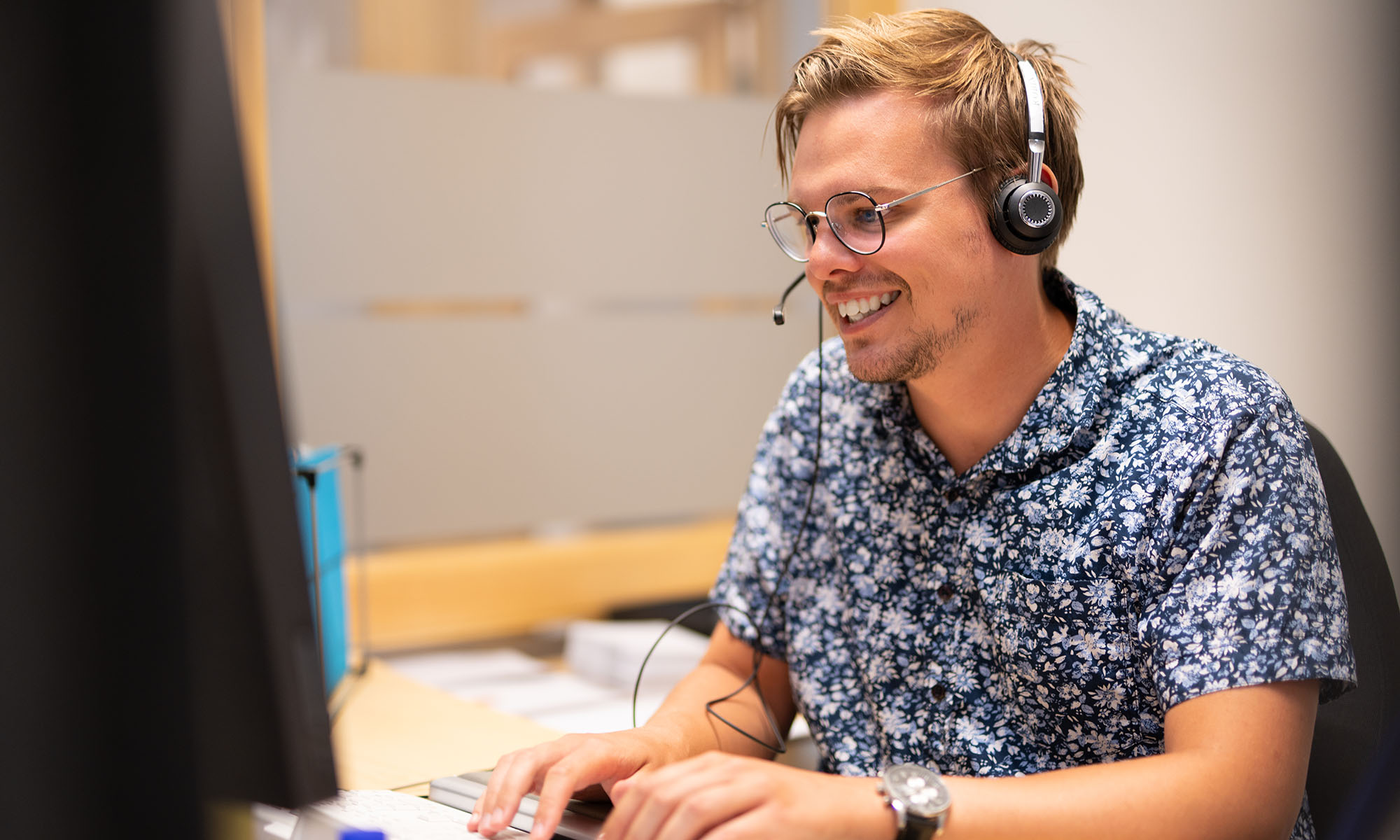 En man med glasögon och en blommig skjorta ler medan han arbetar vid en dator och använder ett headset på ett kontor.